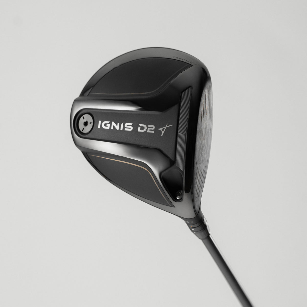 A close-up studio shot of a golf driver head on a light gray background, showing the IGNIS D2 model with a black and dark gray finish and a thin copper accent line.