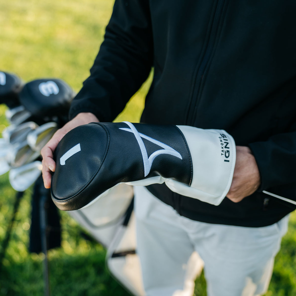 A person outdoors on a grassy golf course holding a black and white Takomo golf club headcover and pulling a driver out.