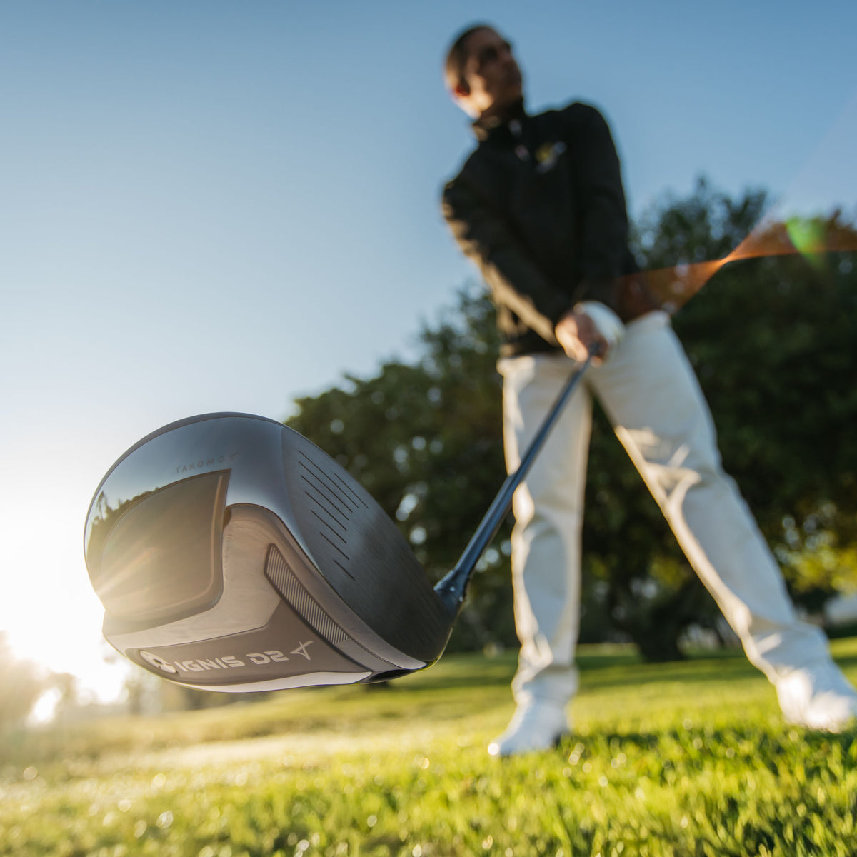 A close-up action shot of a Takomo IGNIS D2 golf driver in the foreground, with a golfer in a black jacket and white pants blurred in the background on a sunny grassy golf course.