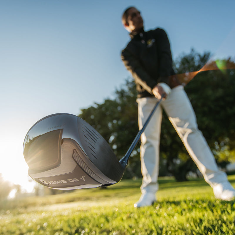 A close-up action shot of a Takomo IGNIS D2 golf driver in the foreground, with a golfer in a black jacket and white pants blurred in the background on a sunny grassy golf course.