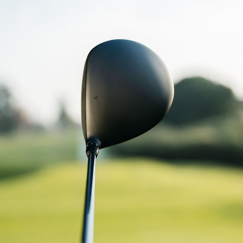 A close-up shot of a matte black IGNIS D2 golf driver head on a blurred green golf course background.