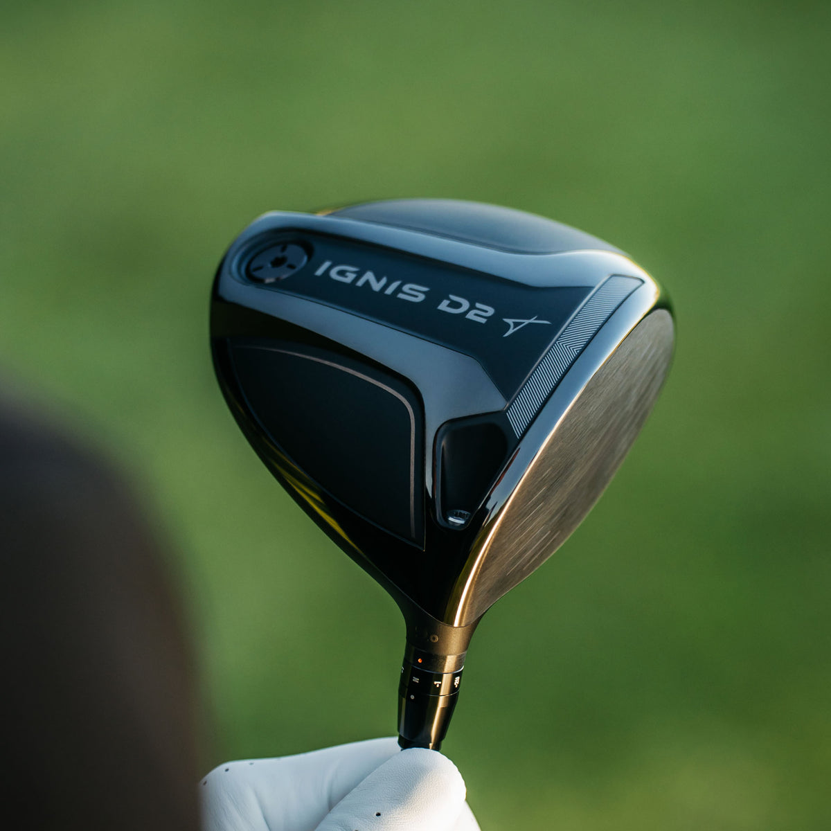 A close-up of a black Takomo IGNIS D2 golf driver head held by a gloved hand against a blurred green grass background.