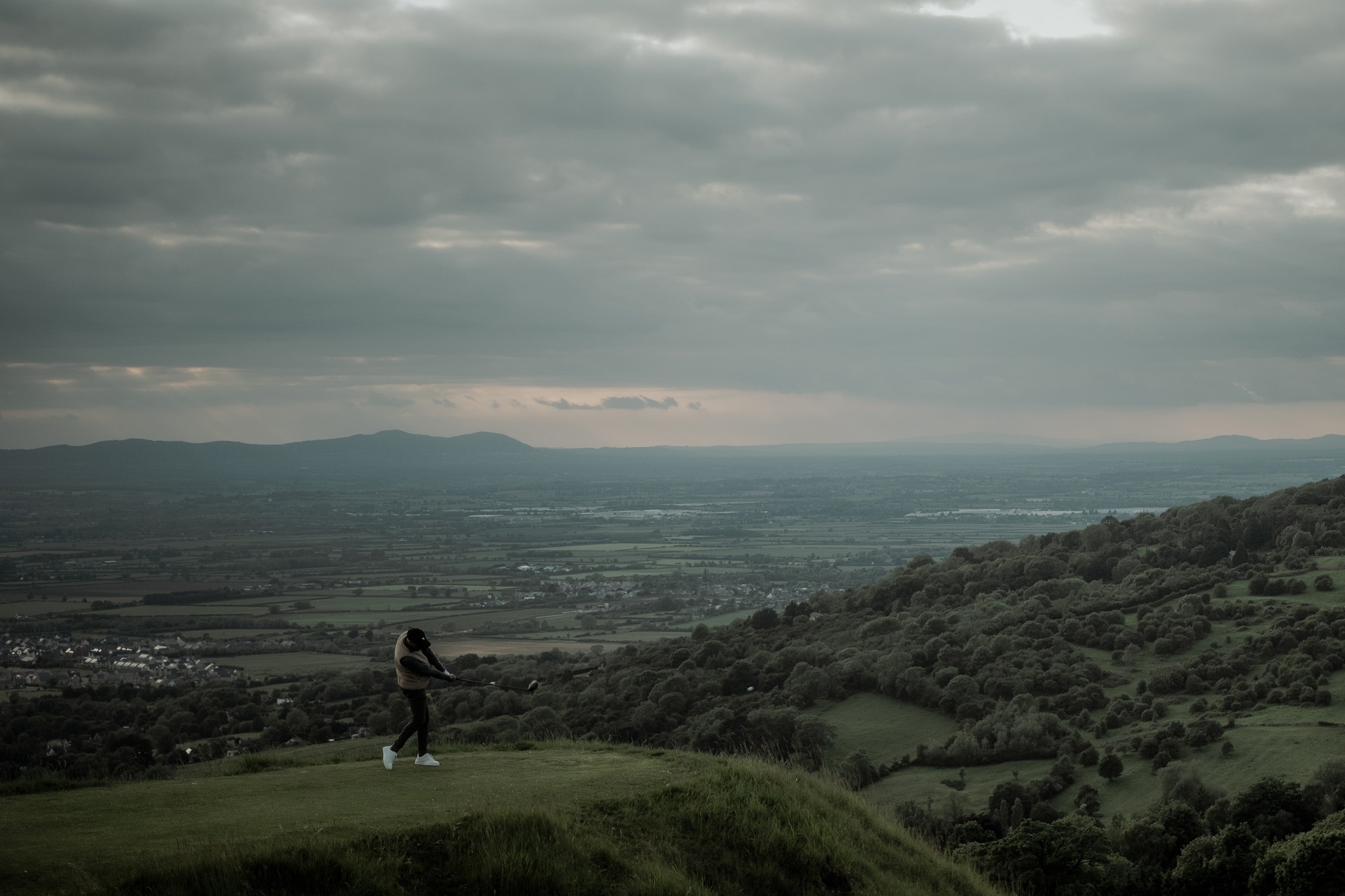 Person golfing on a grassy hill overlooking a vast landscape with a cloudy sky.
