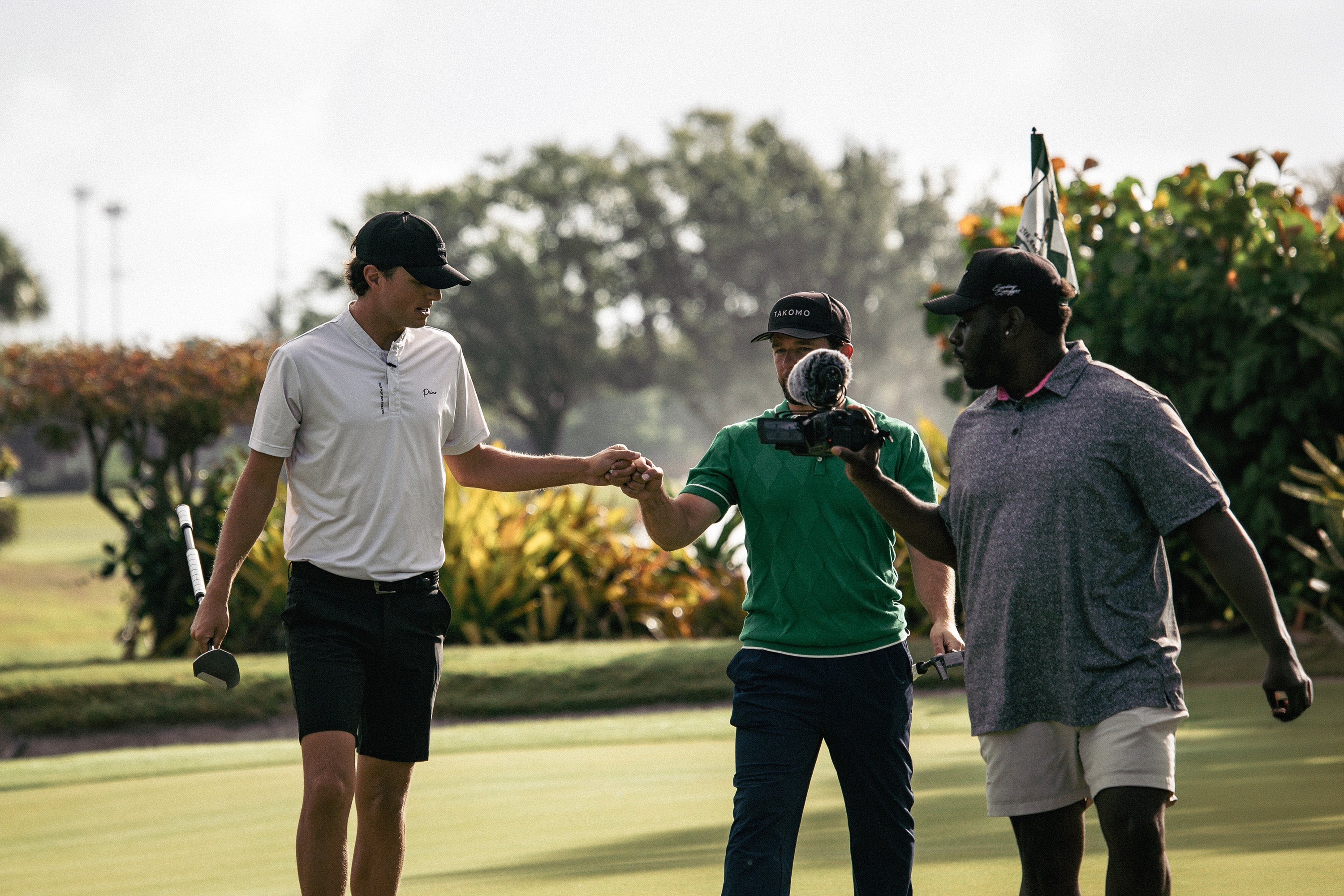 Three men on a golf course with one holding a camera, surrounded by greenery.